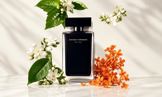 בקבוק בושם Narciso Rodriguez For Her מוקף בפרחים לבנים, עלים ירוקים ופרחים כתומים על רקע בהיר ואלגנטי.
