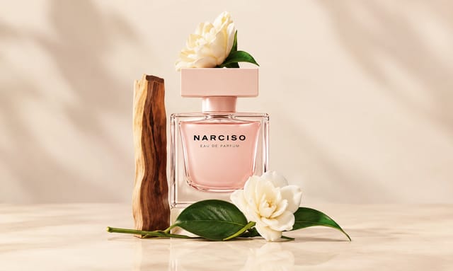 בקבוק בושם NARCISO בגוון ורוד מוקף בפרחים לבנים, עלים ירוקים ואלמנט עץ על רקע בגווני בז׳ רכים.
