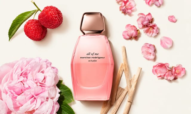 תמונת אווירה של בושם Narciso Rodriguez בגוון ורוד, מוקף בפרחים, פטל ואלמנטים טבעיים על רקע בהיר.