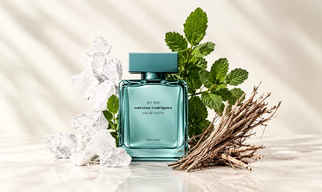 בקבוק בושם Narciso Rodriguez Bleu Noir בגוון טורקיז, מוקף בקרח, עלי מנטה וענפי עץ על רקע בהיר ורך.