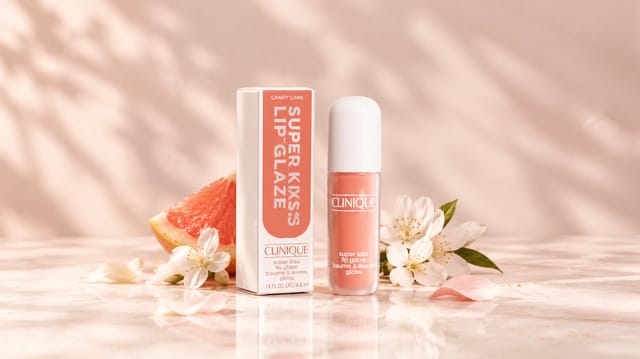 מוצרי Clinique Super Kiss Lip Glaze בגוון ורוד־קורל, לצד אשכולית ופרחים לבנים על רקע ורוד־אפרסק רך.