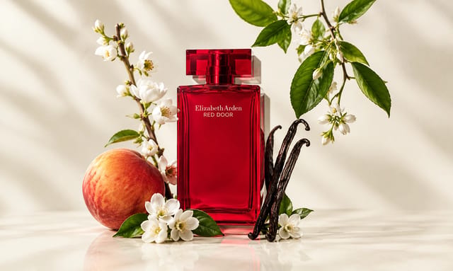 בקבוק בושם Elizabeth Arden Red Door בגוון אדום עמוק, מוקף בפרחים לבנים, עלים ירוקים, אפרסק ותרמילי וניל על רקע בהיר.