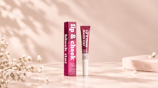 מוצרי Lip & Cheek בגוון ורוד־בורדו על רקע ורוד בהיר, לצד פרחים לבנים ואלמנטים עדינים.