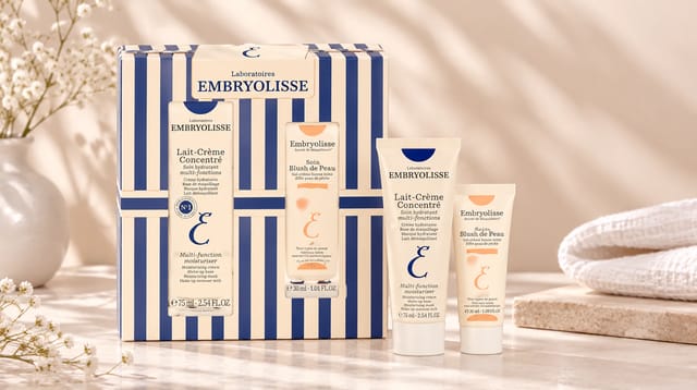 מארז טיפוח ומוצרי פנים של המותג Embryolisse, על רקע בגוון בז' עדין עם פרחים לבנים ומגבת רכה.