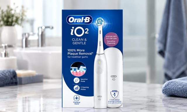 מברשת השיניים החשמלית Oral-B iO² Clean & Gentle מוצגת על משטח שיש בחדר אמבטיה מואר, עם כיור, ברזים ומגבות ברקע מטושטש.